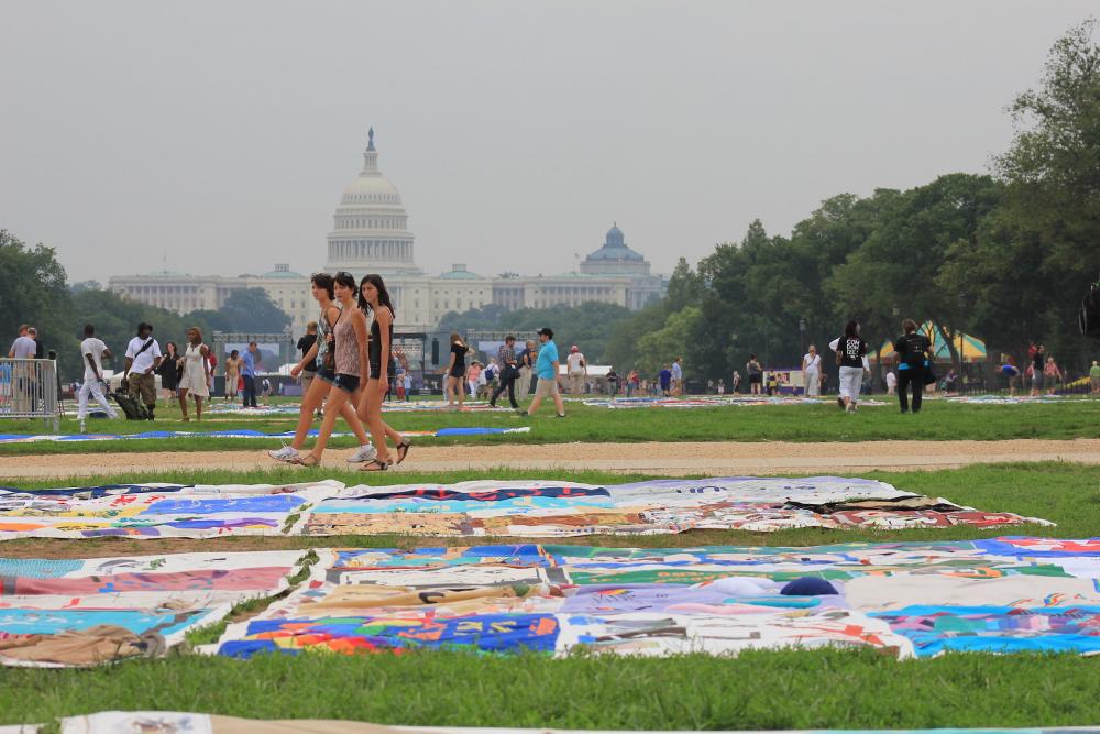 three people walking on sidewalk with Capitol building in background, looking at the AIDS quilt on the National Mall