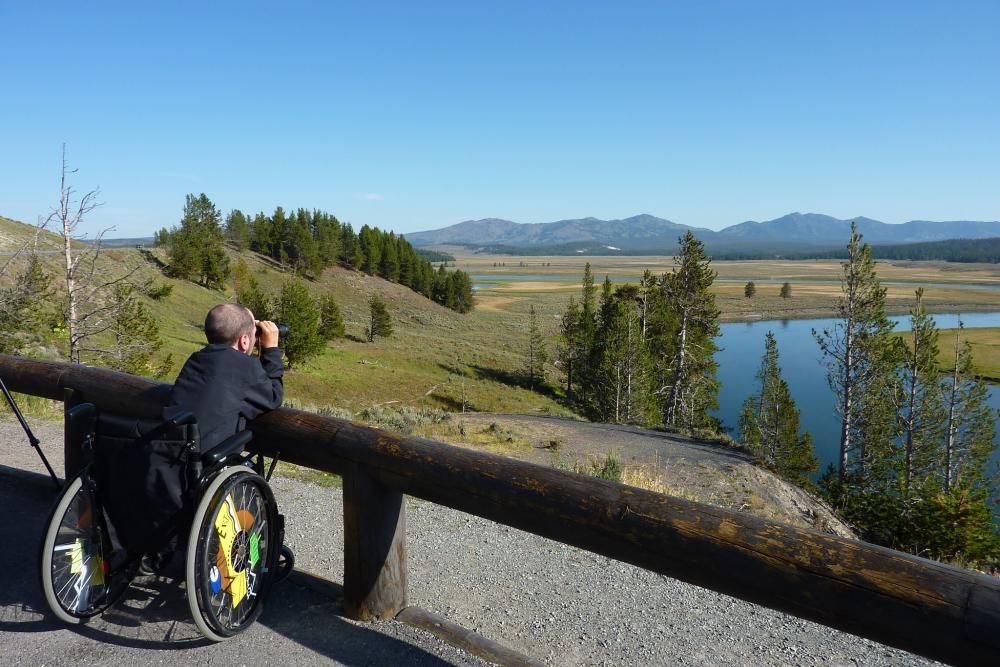 Person sitting in wheelchair looking through binoculars at wide-open scenic vista 