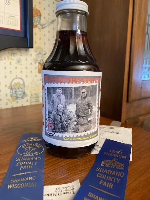 A bottle of Miller Brothers Sugar Bush maple syrup sits on a desk with two first place ribbons next to them.