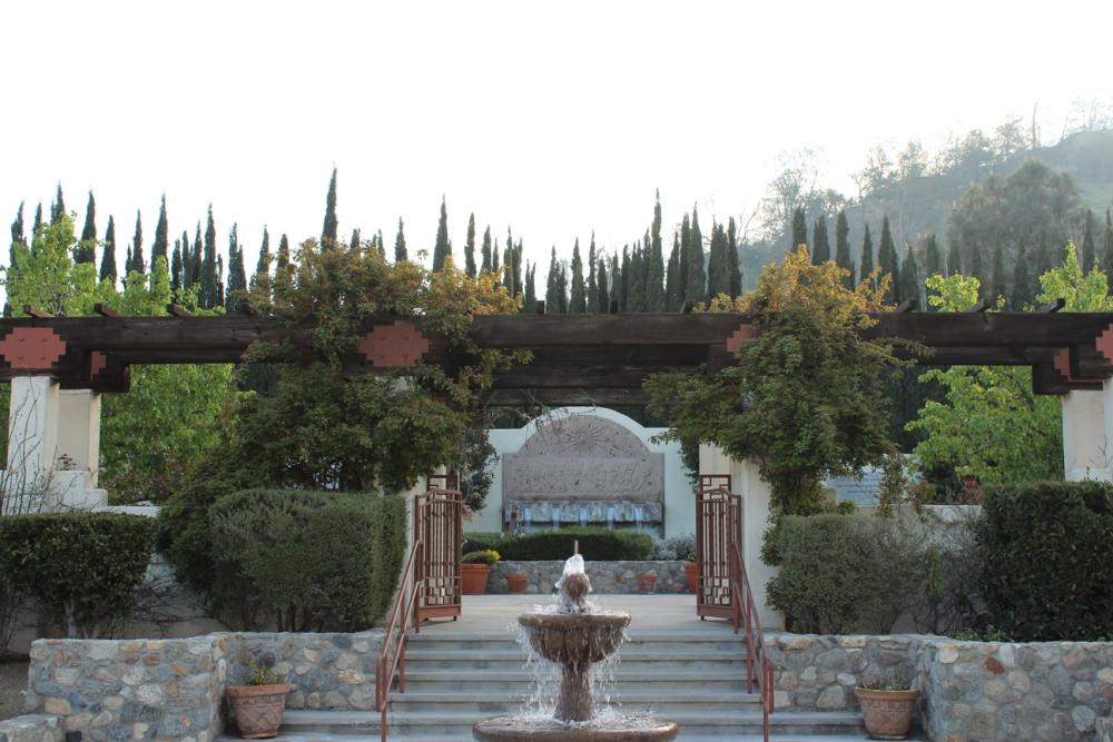 A fountain flows in front of a set of stairs that lead to a path that passes below a wooden arbor. In the background is a rectangular fountain with an arched top