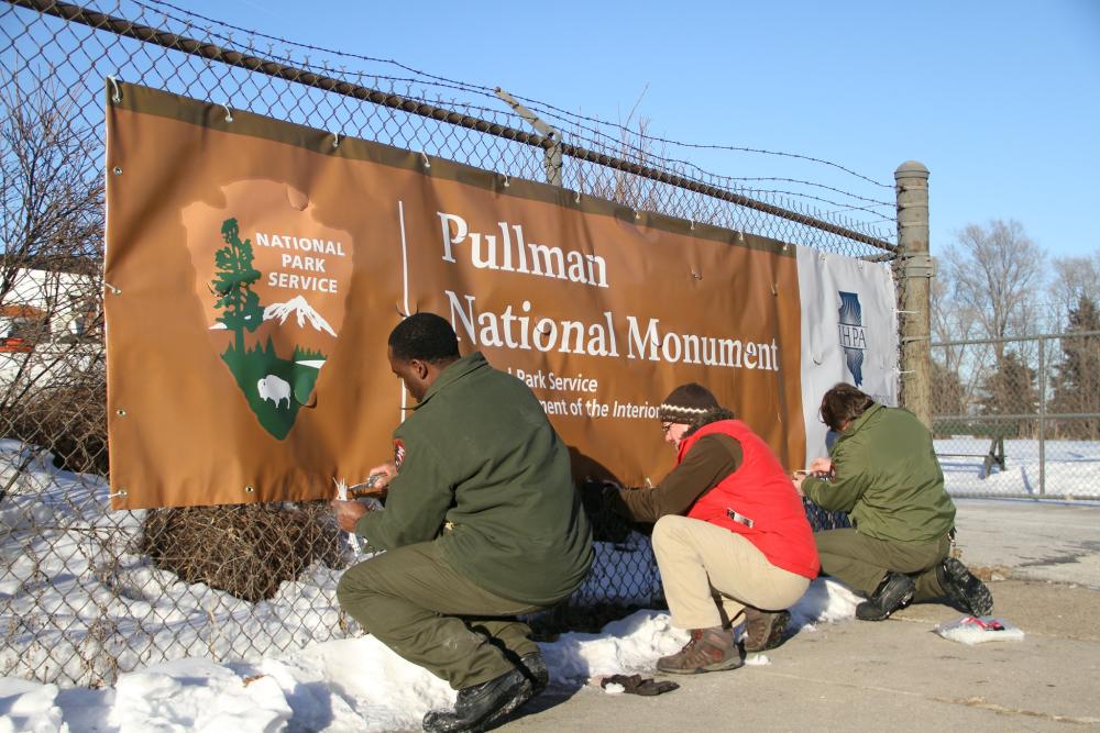 Three people kneel while securing a banner that reads "Pullman National Monument" against a chain-link fence