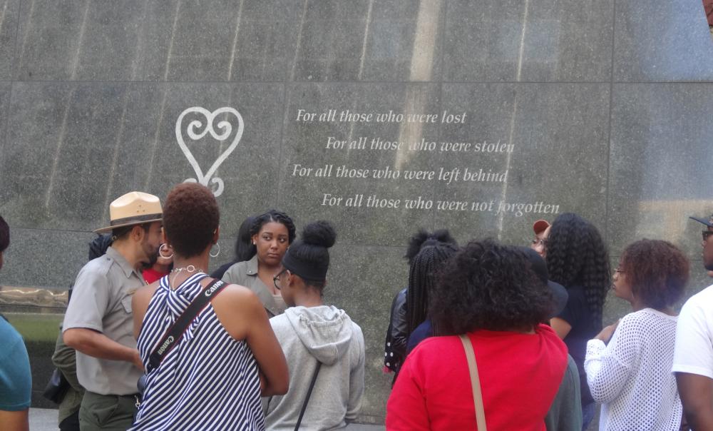 Group of people in conversation standing in front of a dark stone polished surface on which these words are visible: "For all those who were lost/For all those who were stolen/For all those who were left behind/For all those who were not forgotten"