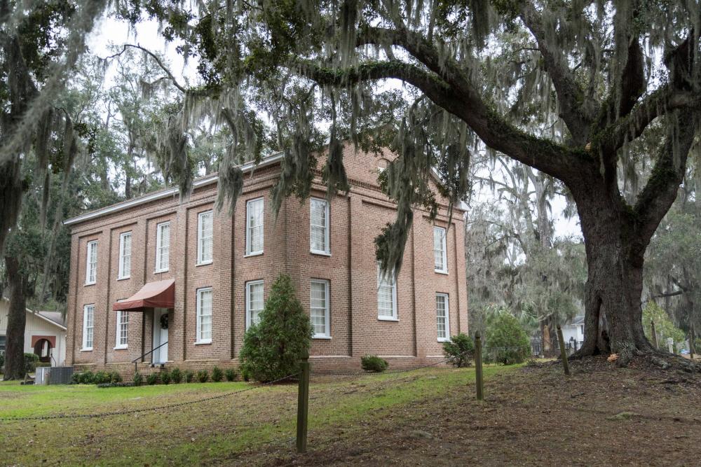 Brick building with many windows and small reddish awning atop front door. Spanish moss is visible hanging from tree branches in foreground and background.